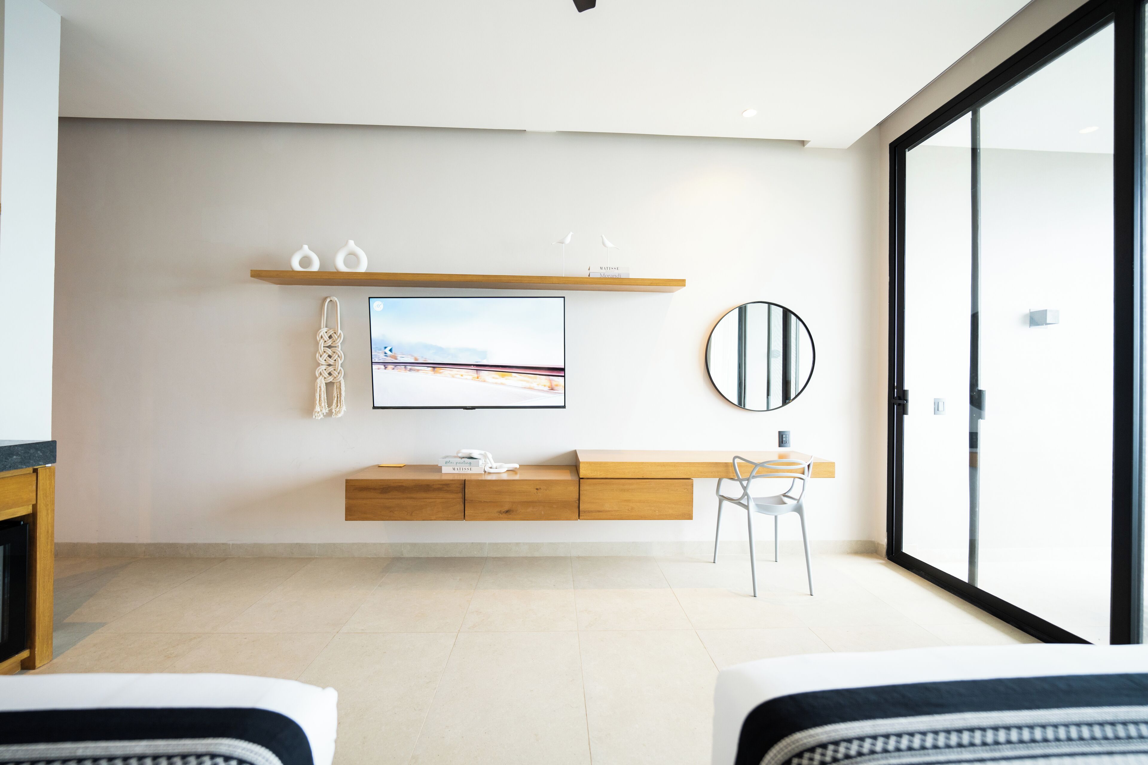 Quarto de hotel moderno com uma janela grande, televisão de tela plana na parede, mesa de console de madeira, espelho redondo e um espaço de estar com duas cadeiras.