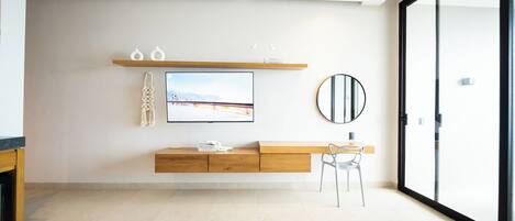 Quarto de hotel moderno com uma janela grande, televisão de tela plana na parede, mesa de console de madeira, espelho redondo e um espaço de estar com duas cadeiras.