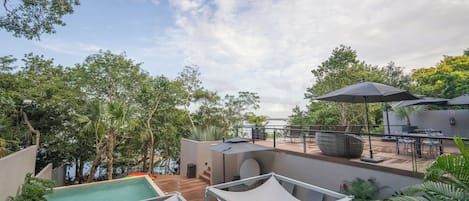 A rooftop pool area with lounge chairs and umbrellas, surrounded by trees and overlooking a body of water.