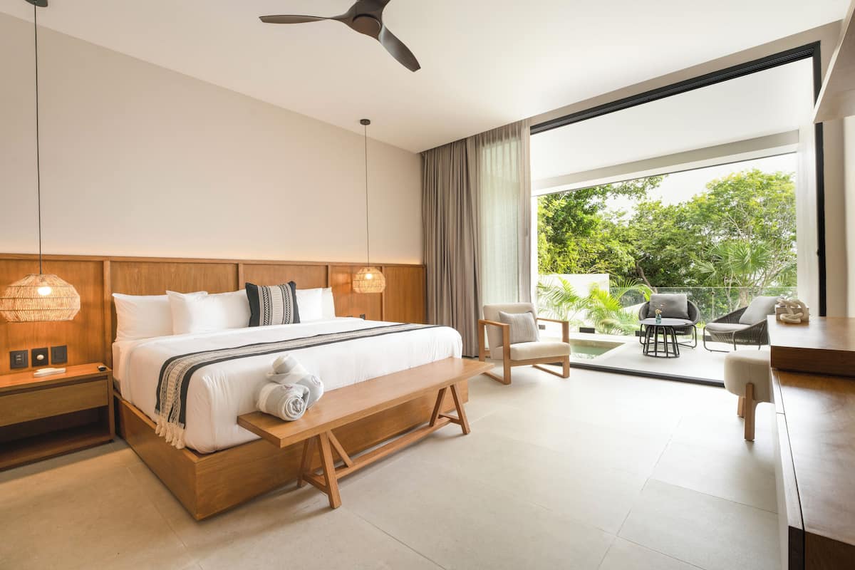 A modern hotel room with a large bed, wooden headboard, bedside tables, a bench, a desk, and a view of the outdoors.