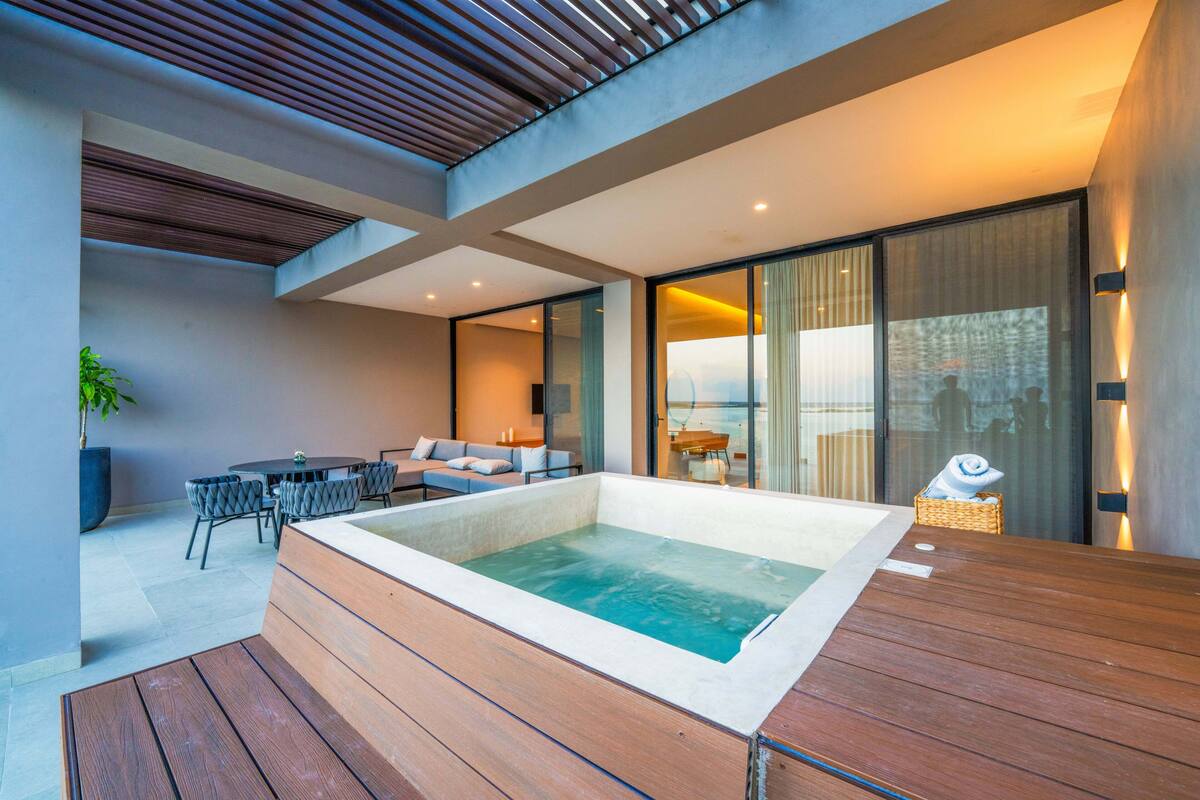 A modern outdoor pool area with a wooden deck, a seating area with a sofa and chairs, and a large window offering a view of the city.