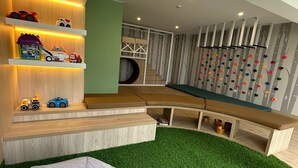 Un salon moderne avec un tapis vert en gazon synthétique, un canapé blanc, des étagères en bois remplies de jouets et un mur d’escalade.
