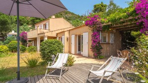 Une terrasse en bois avec deux chaises longues blanches, un grand parasol et une maison à la façade beige et aux volets blancs.