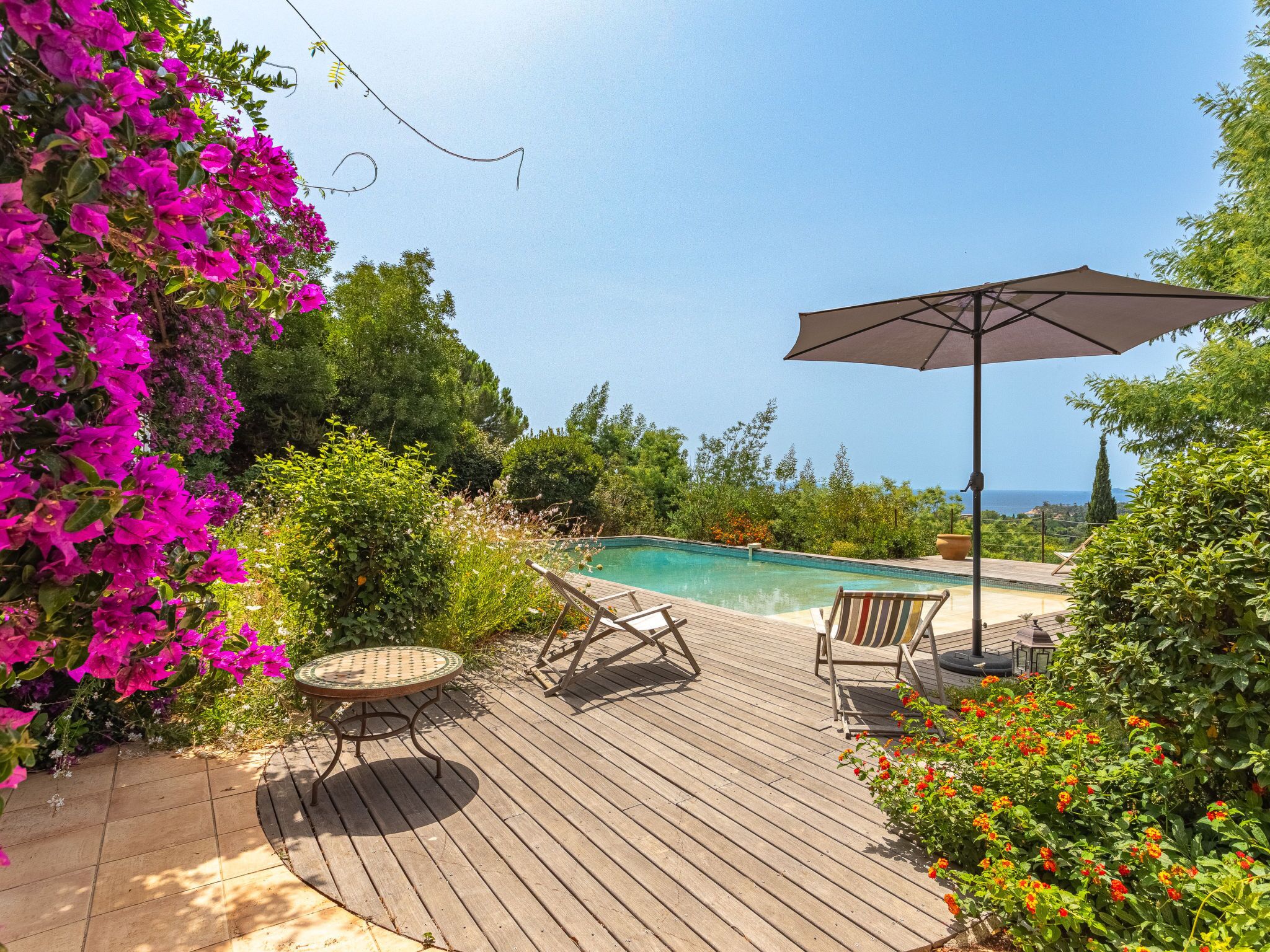 Una terrazza in legno con piscina, circondata da vegetazione rigogliosa e fiori colorati.