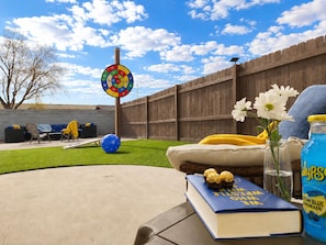 A backyard with a wooden fence, a dartboard, a blue ball, a cushion, a book, a vase with flowers, and a lemonade bottle.