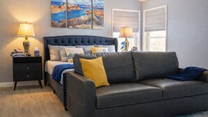 A bedroom with a dark-colored sofa, a bed with multiple pillows, a nightstand with a lamp, and a painting above the bed. - Big house w custom murals/2 Kings/Fun backyard w Games/Grill/Dog OK/Boat parking (Page)