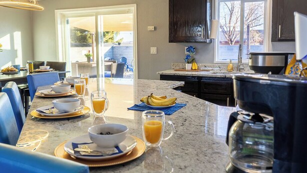 A modern kitchen with granite countertops, a coffee maker, and a dining area with blue chairs and a table set for breakfast. - Big house w custom murals/2 Kings/Fun backyard w Games/Grill/Dog OK/Boat parking (Page)