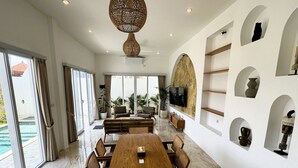 A spacious living area with a large wooden dining table, comfortable seating, a television mounted on a stone wall, and a view of the outdoor swimming pool. - Villa Amici (Canggu)