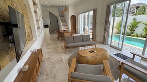 A modern living room with a TV, wooden furniture, a round coffee table, and a view of a swimming pool. - Villa Amici (Canggu)