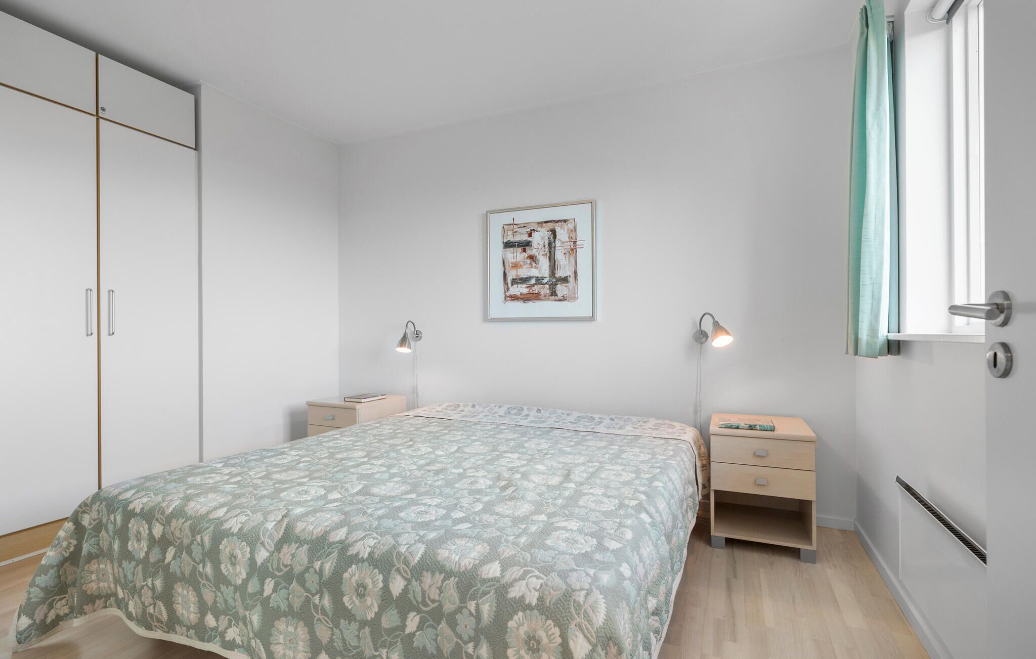 A bedroom with a bed, two bedside tables, a wardrobe, a framed picture on the wall, and a window with curtains.