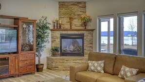A living room with a stone fireplace, a television, a sofa, and a view of a lake. - Lakefront, Fireplace, Large Deck, Yard & Dock. Mins from Uptown Dining & Trails! (Pagosa Springs)