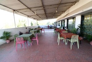 Un espace extérieur aménagé en terrasse, avec des tables et des chaises, recouvert d’un toit en métal, et agrémenté de plantes en pot.