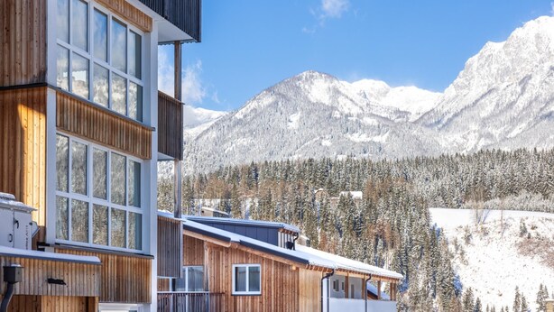 A modern building with large windows and a wooden facade, set against a backdrop of snow-covered mountains and a clear blue sky. - Atmospheric with a sunny balcony and summercard (Haus im Ennstal)