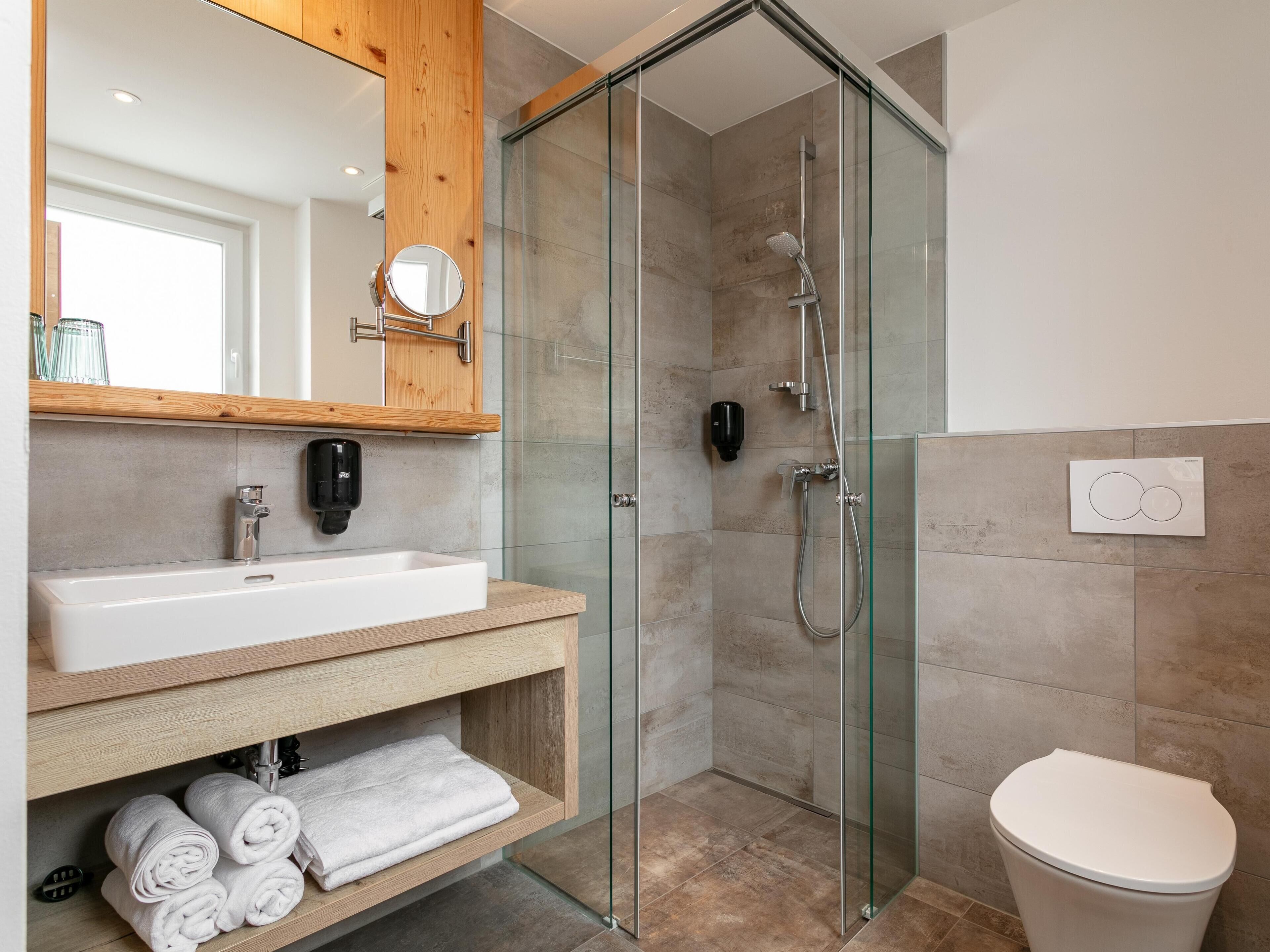 Un baño moderno con ducha envuelta en vidrio, lavamanos blanco y tocador de madera.