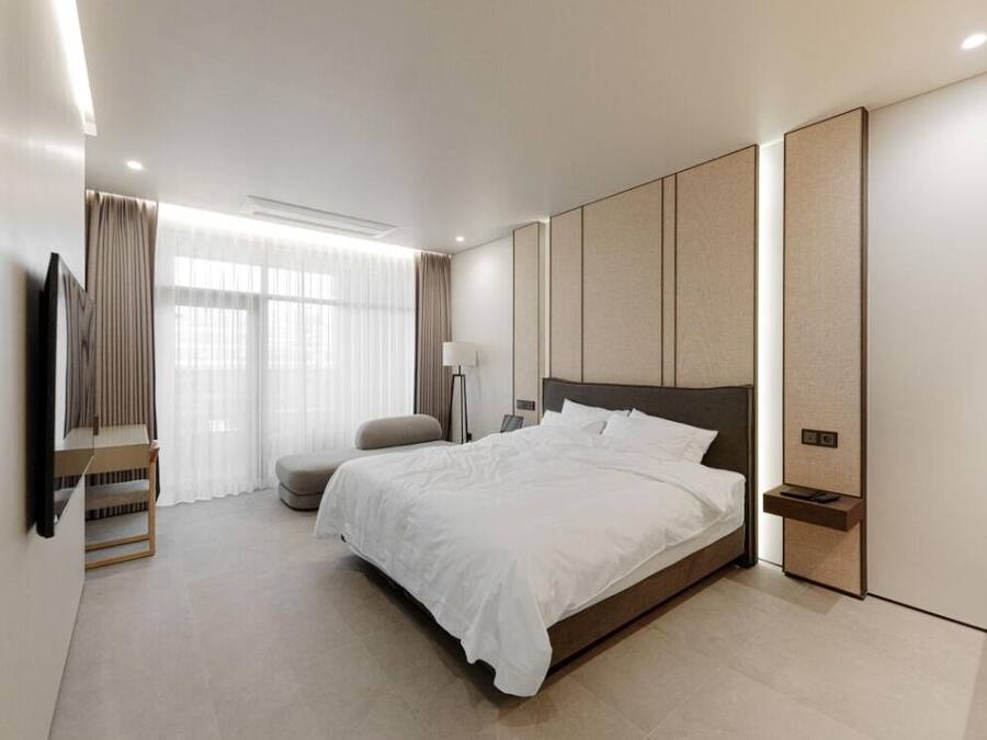 A modern hotel room with a large bed, a flat-screen TV mounted on the wall, a bedside table, and a large window with curtains.