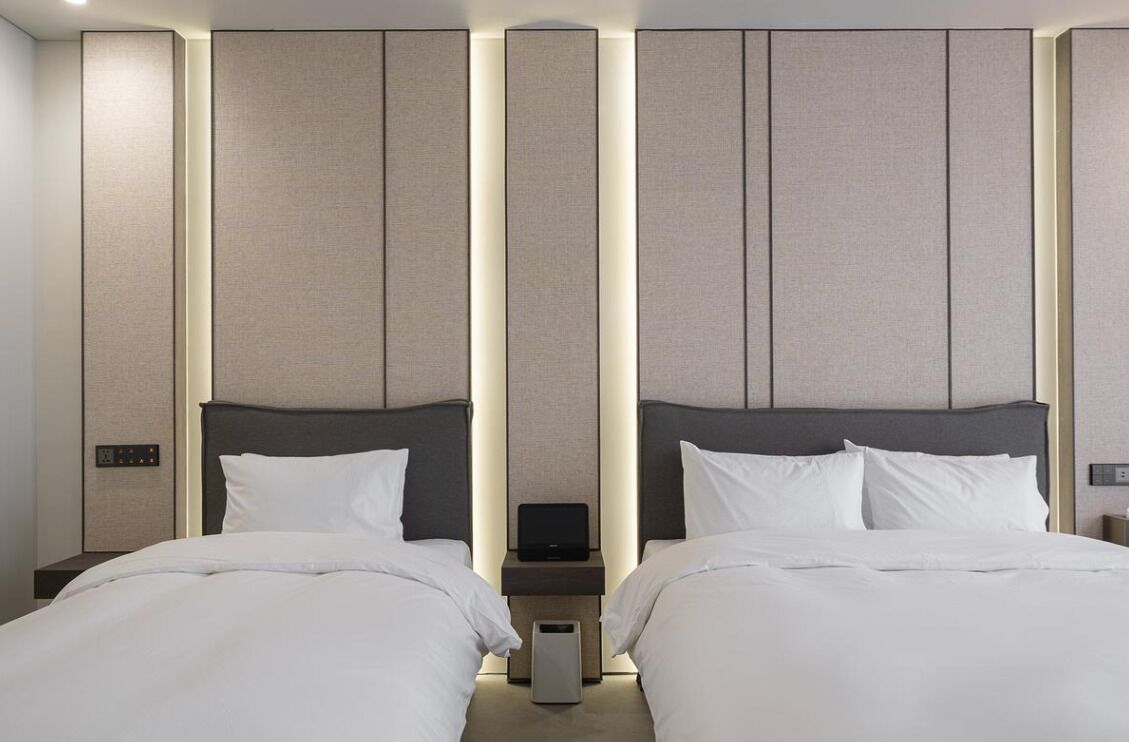 A hotel room with two beds, a bedside table with a lamp, and a wall panel design.