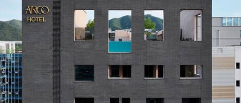 Un bâtiment moderne dont la façade porte l’inscription « ARCO HOTEL ». Il est doté de nombreuses fenêtres, dont certaines offrent une vue sur la verdure et une piscine.