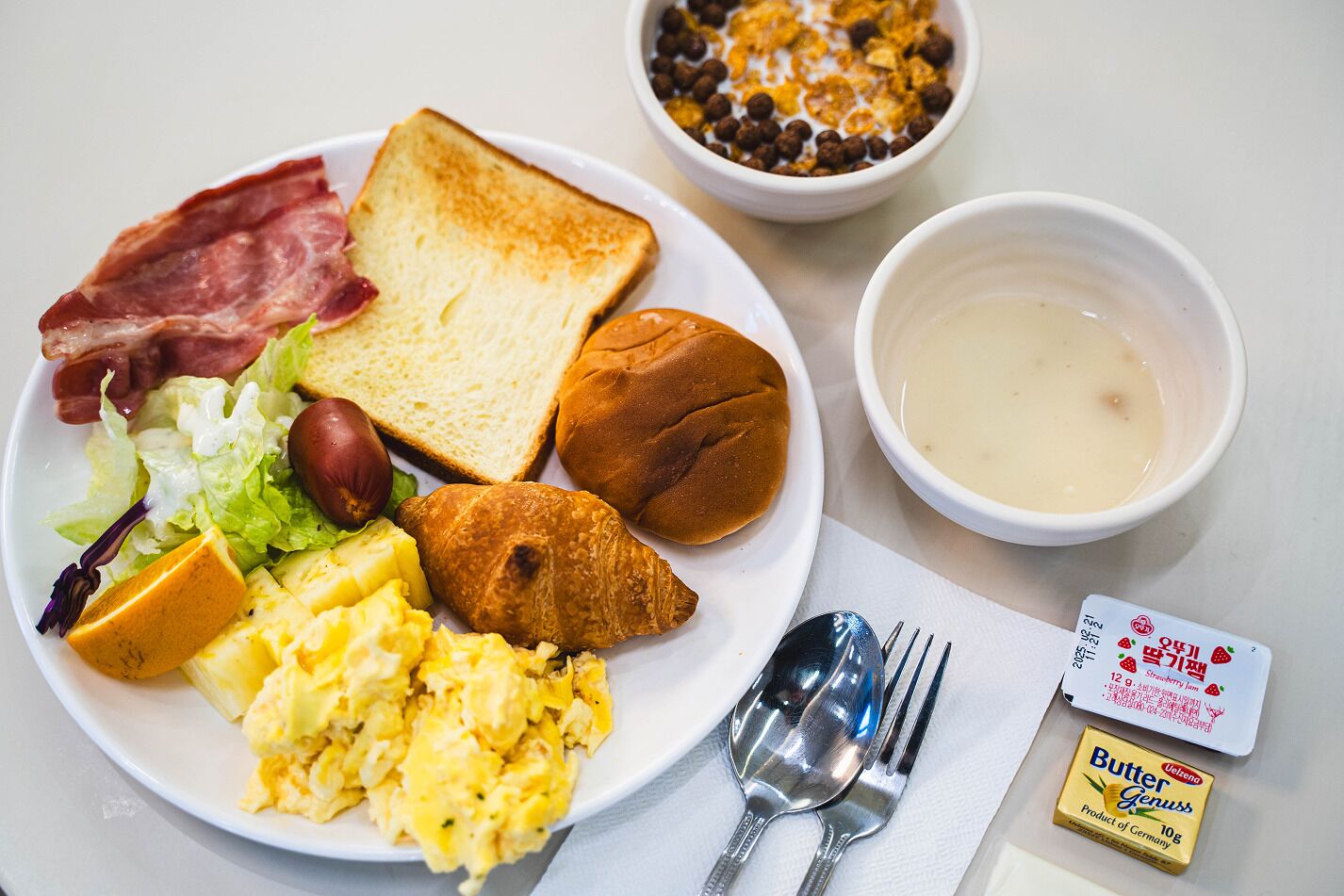 Un plato de desayuno con tostadas, salchicha, huevos, un croissant y un bol de sopa.