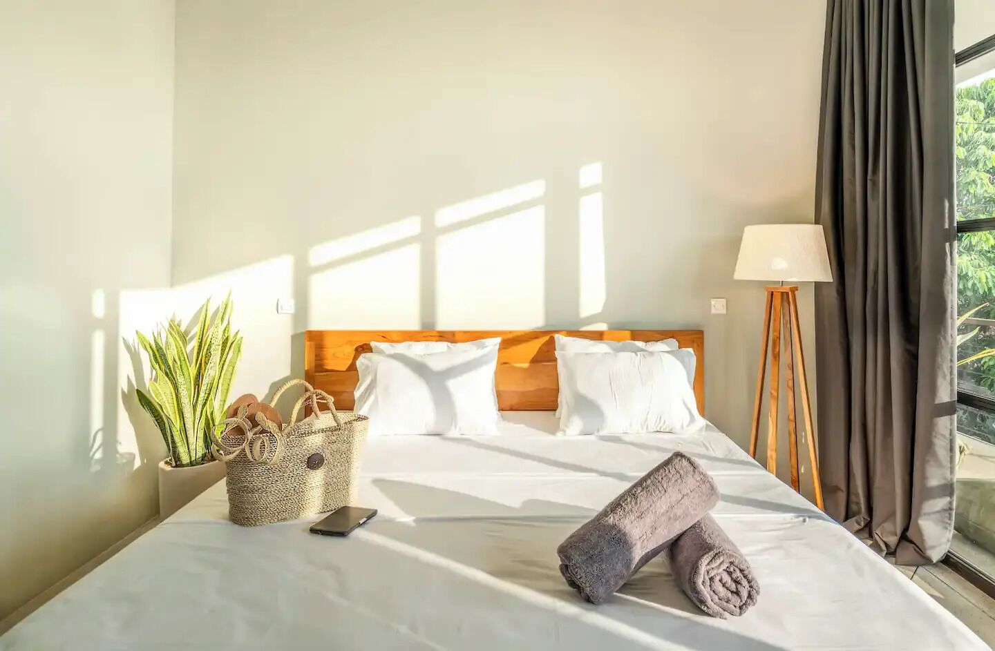 A hotel room with a bed, a potted plant, a woven basket, and a lamp.