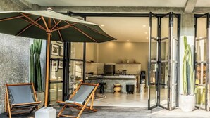 A modern kitchen with a central island, a dining area with two chairs and an umbrella, and a view of the living area through glass doors. - Lane House 2 (Canggu)