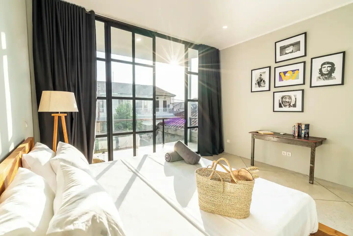 A hotel room with a large window, a bed, a bedside lamp, a small table with books, and framed pictures on the wall.