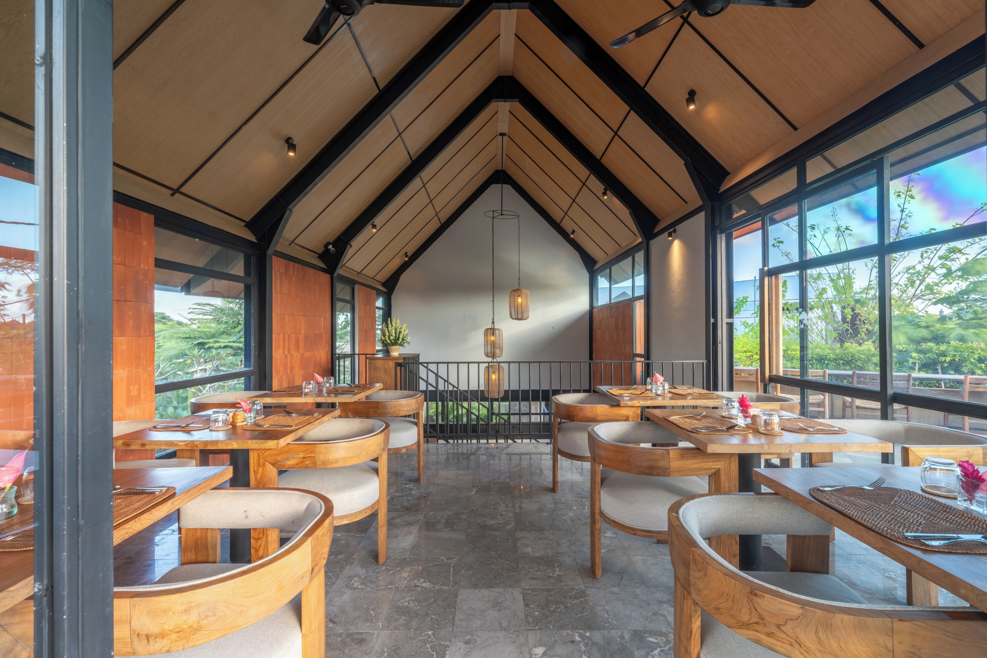 Un restaurante con mesas y sillas de madera, ventanas amplias y vistas a zonas verdes.