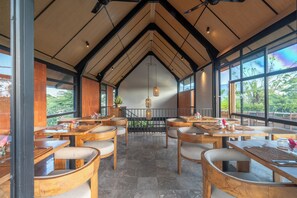Un restaurant avec des tables et des chaises en bois, de grandes fenêtres et une vue sur la verdure environnante.