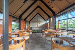 A restaurant with wooden tables and chairs, large windows, and a view of greenery outside.