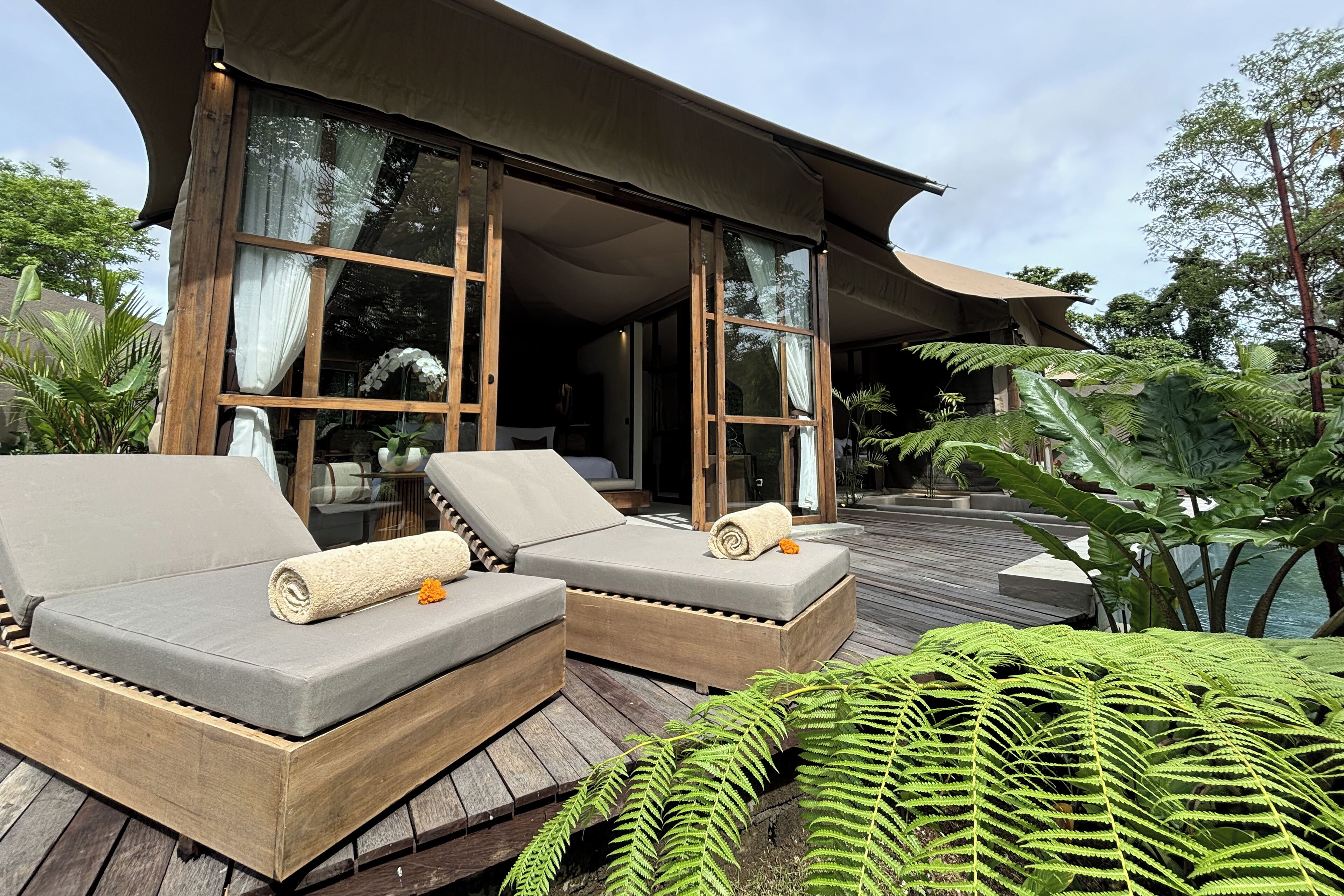 A wooden deck with two grey lounge chairs, a large window, and lush greenery.