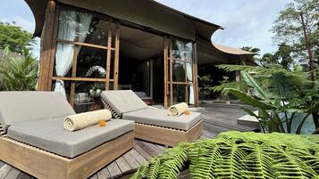 A wooden deck with two grey lounge chairs, a large window, and lush greenery.