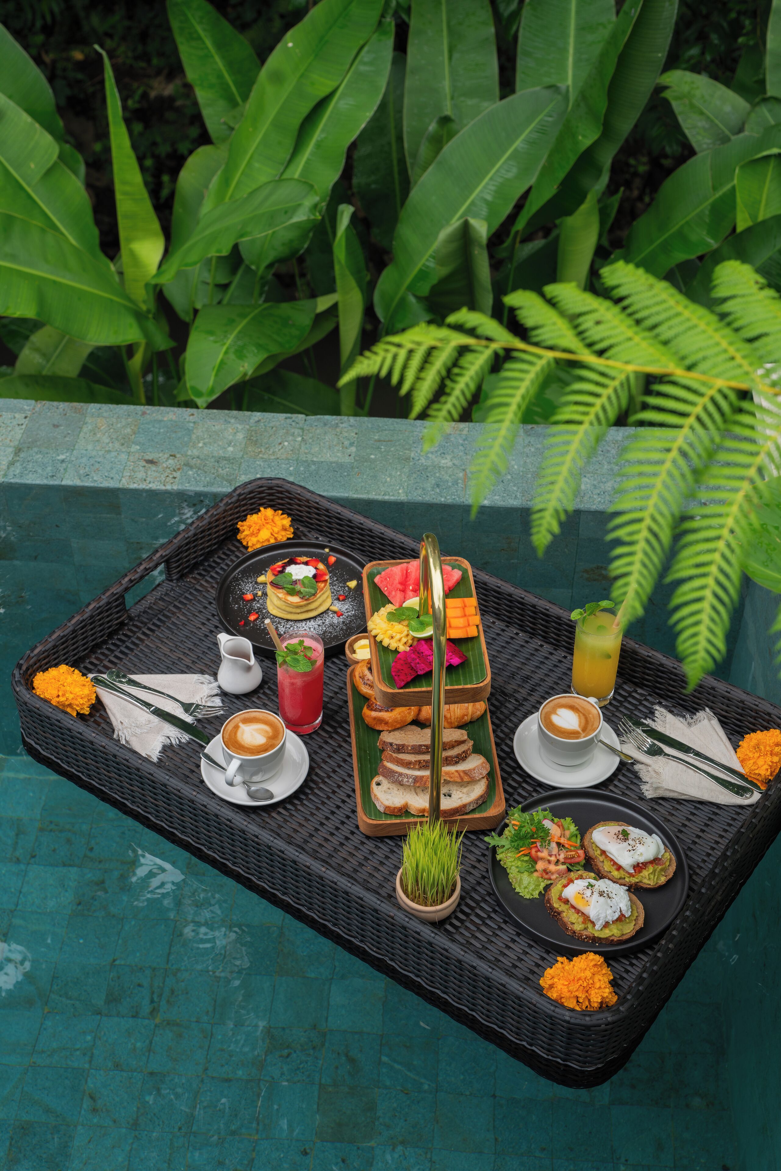 Uma bandeja de vime com itens de café da manhã, incluindo torradas, ovos e uma bebida, colocada em uma mesa à beira da piscina com vista para vegetação exuberante.