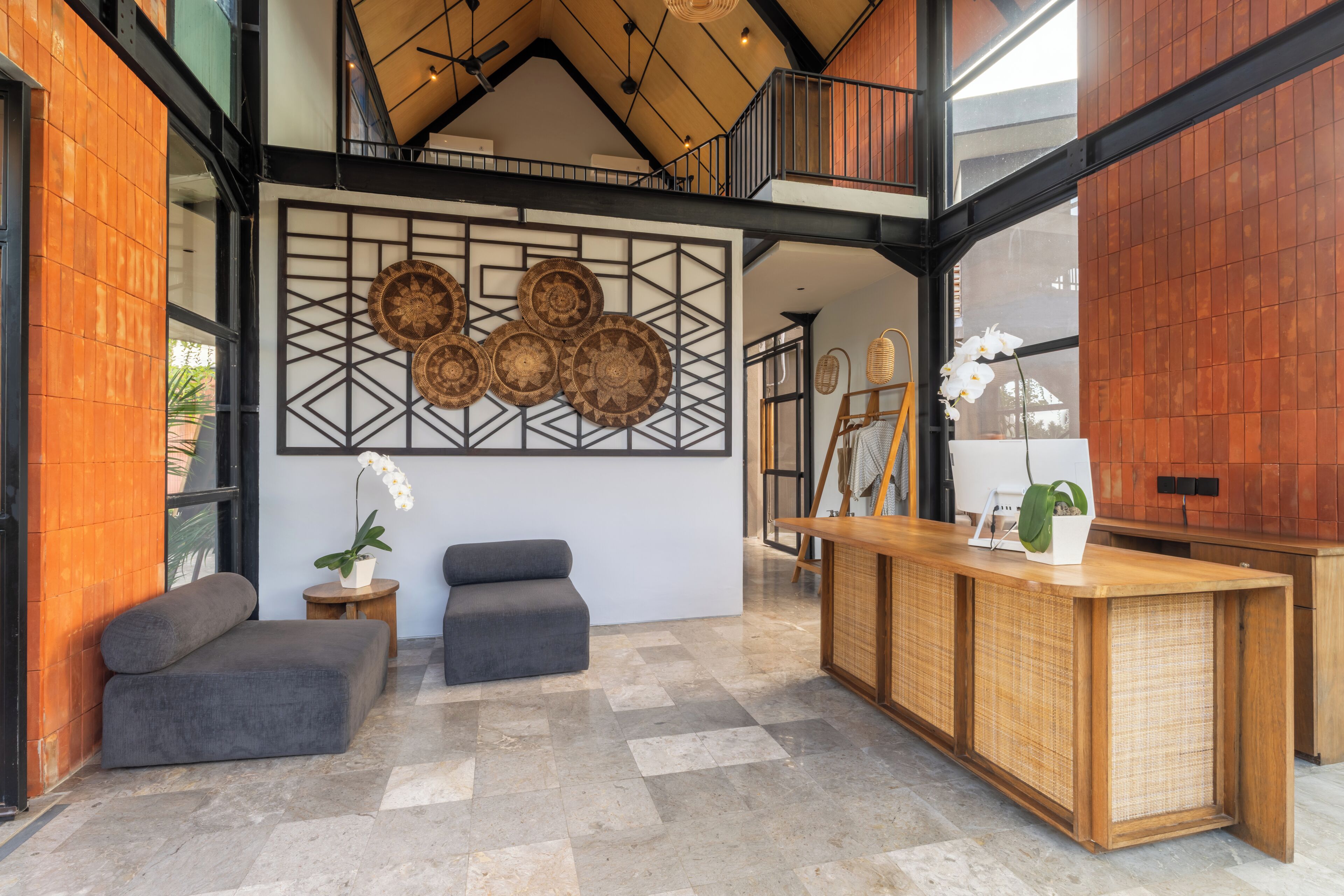 Un área de recepción con techo alto, mostrador de madera, dos sofás grises y una pared decorativa con diseños tejidos.