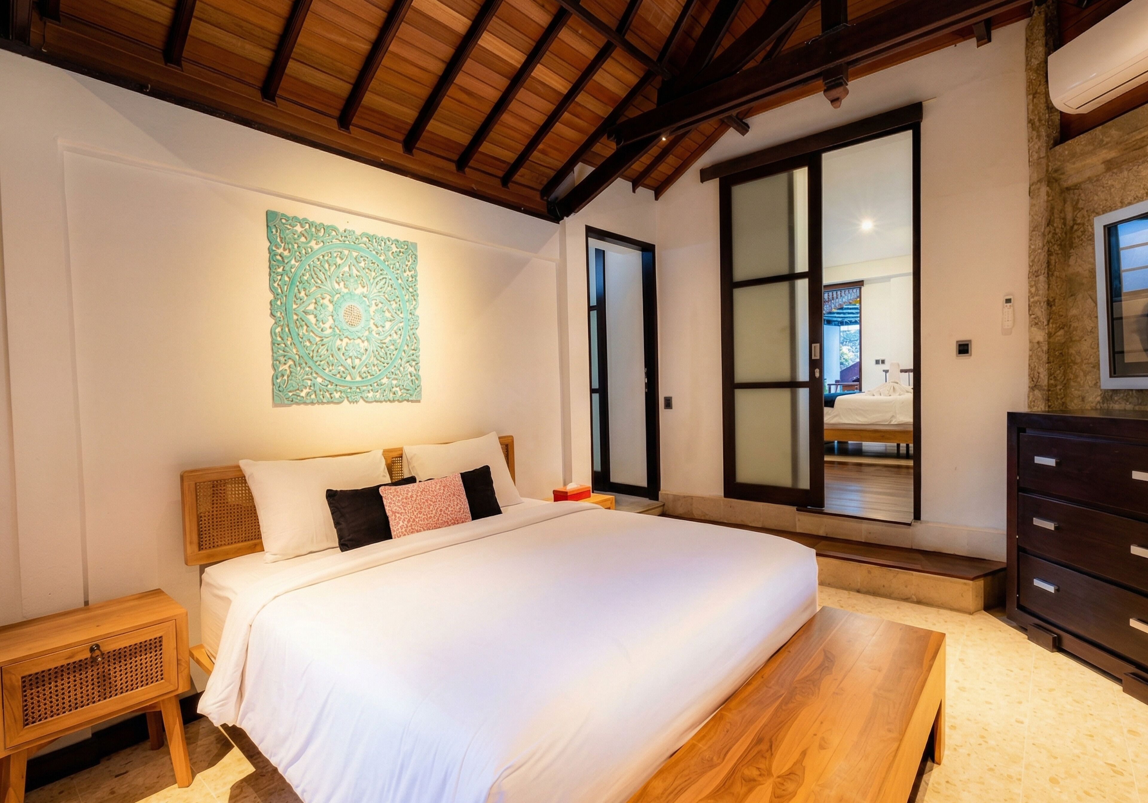 A bedroom with a large bed, wooden headboard, a nightstand, a wooden bench, a dresser, and a large window with a view.