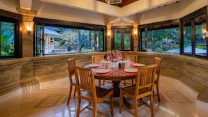 A dining area with a round table, wooden chairs, and a view of greenery outside. - Villa Frangipani Seminyak (Seminyak)