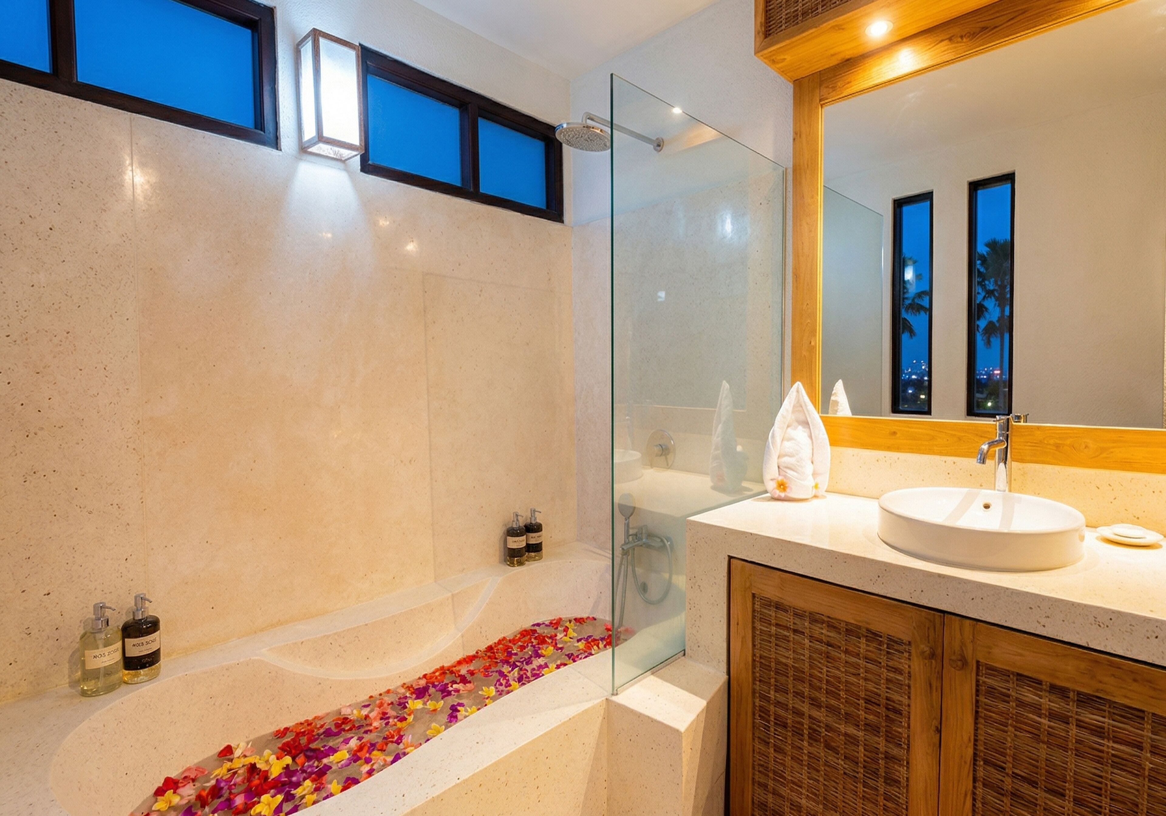 Un baño moderno con ducha de vidrio, una bañera con pétalos de rosa, un armario de madera y un espejo grande.