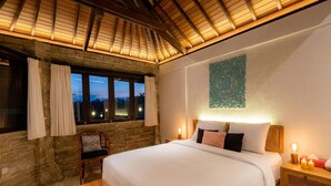 A bedroom with a large bed, a wooden headboard, a nightstand with candles, a chair, and a window with curtains. - Villa Frangipani Seminyak (Seminyak)