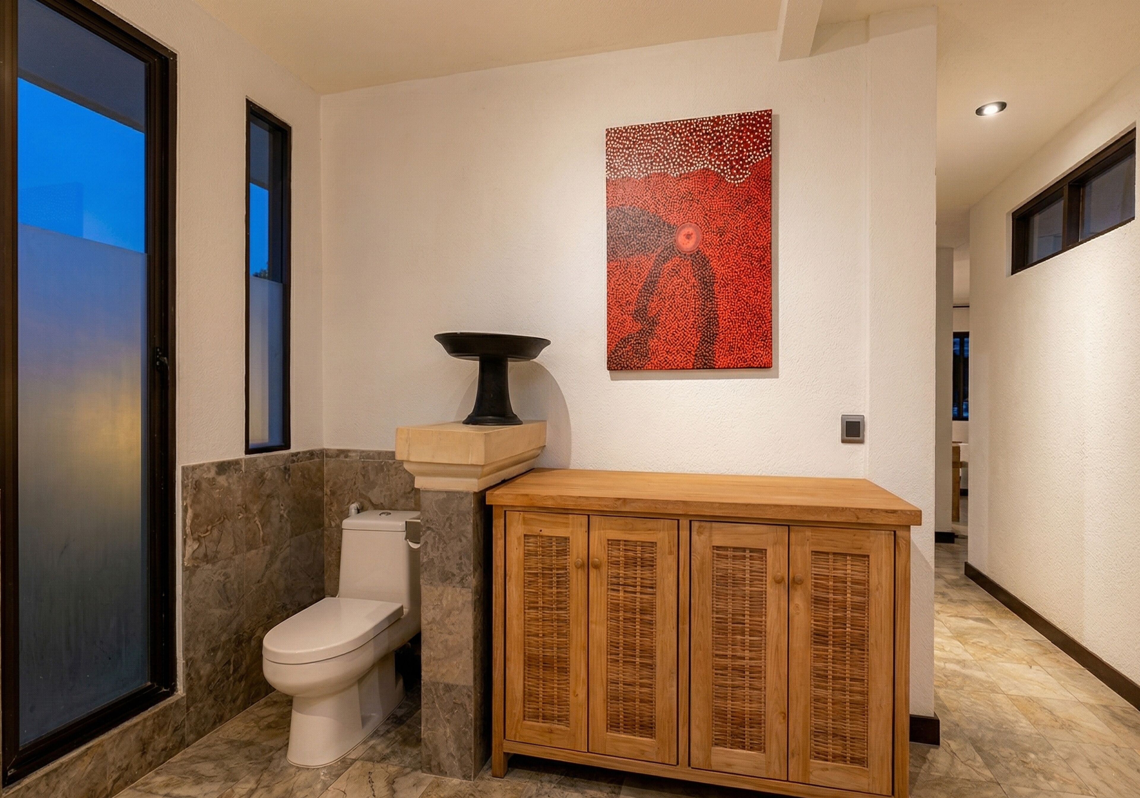 A bathroom with a toilet, a wooden cabinet, and a wall-mounted artwork.
