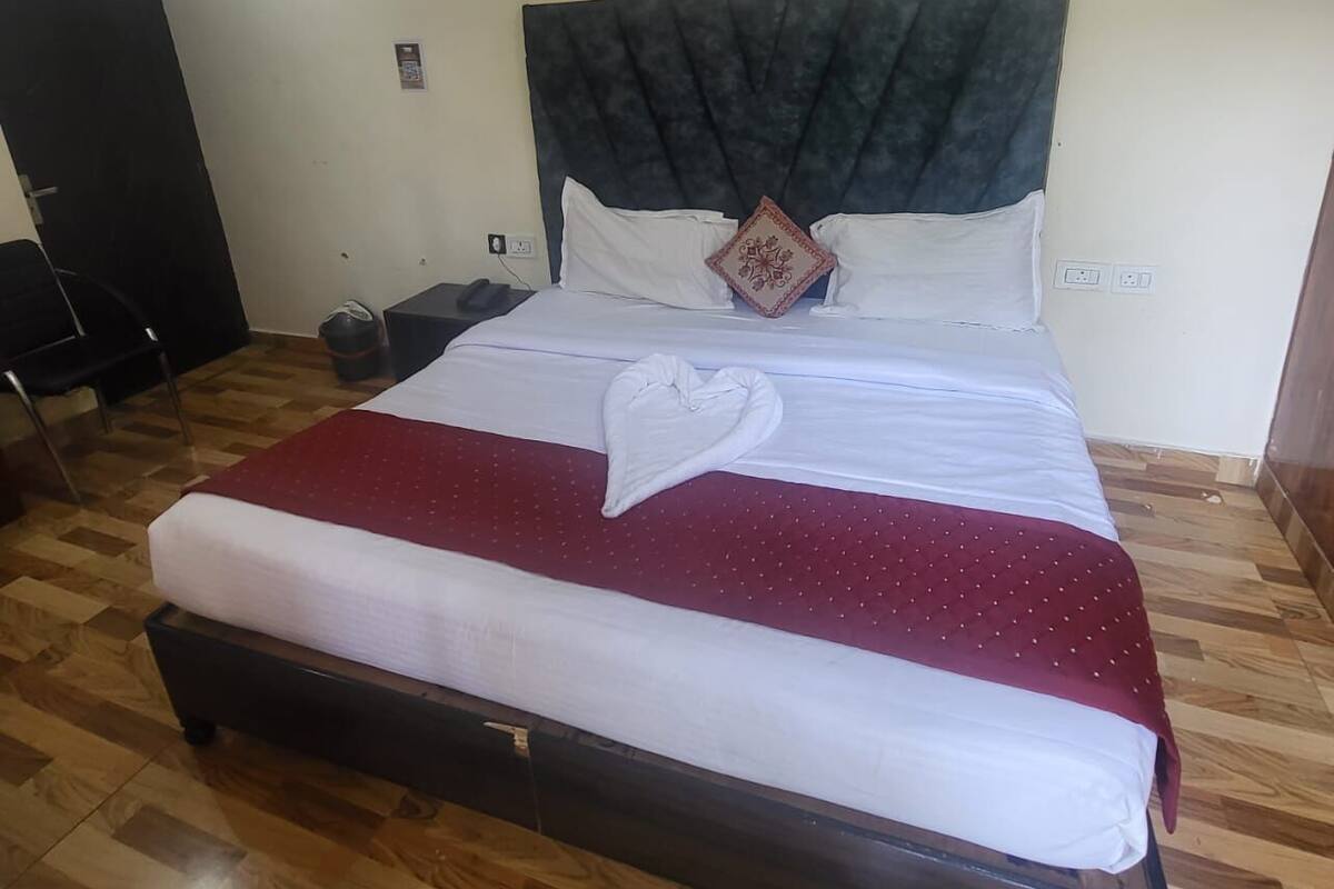 Un lit bien fait, avec une tête de lit sombre, du linge de lit blanc et une couverture rouge. Un petit coussin décoratif est posé sur le lit. La pièce est dotée d'un parquet et d'un meuble noir situé à gauche.
