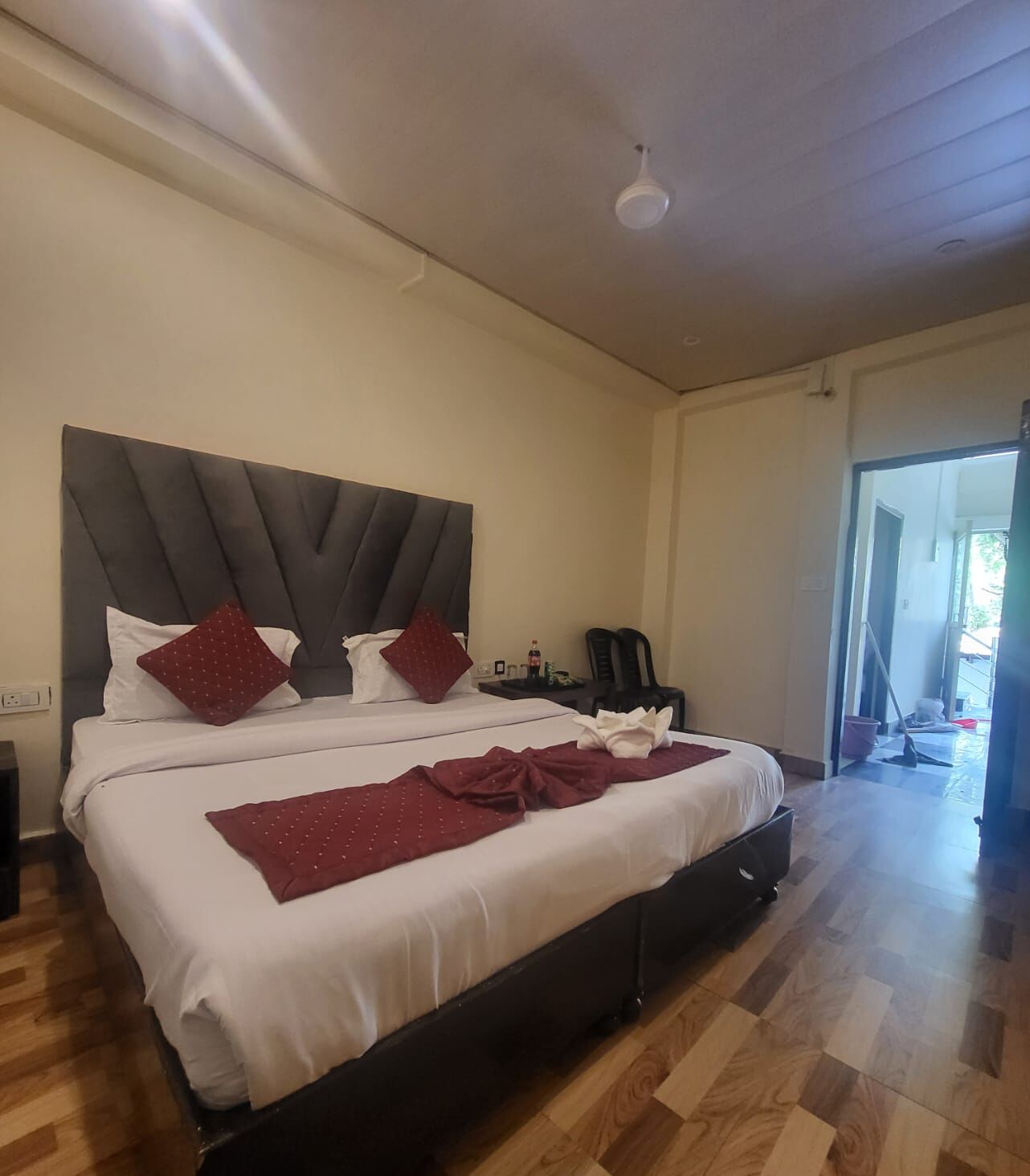 Una cama grande con cabecera oscura, ropa de cama blanca y almohadas rojas. El suelo es de madera y se ve una puerta que da a otra habitación.