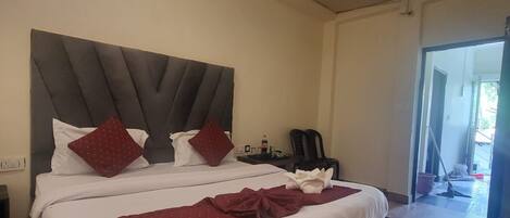 Una cama grande con cabecera oscura, ropa de cama blanca y almohadas rojas. El suelo es de madera y se ve una puerta que da a otra habitación.