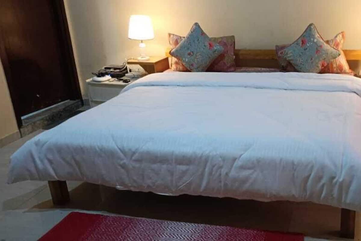 Cama bem arrumada com lençóis brancos e almofadas decorativas, duas luminárias de cabeceira e um tapete vermelho no chão.