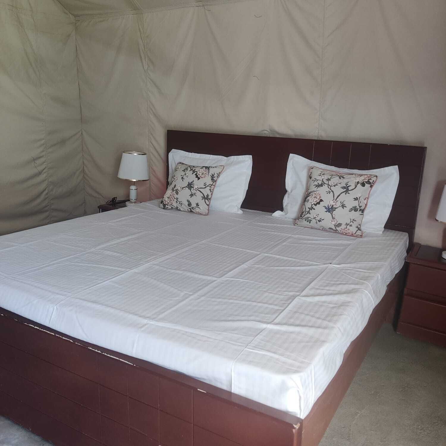 Una cama con colcha blanca, dos almohadas florales, un cabecero de madera y una mesita de noche con lámpara.