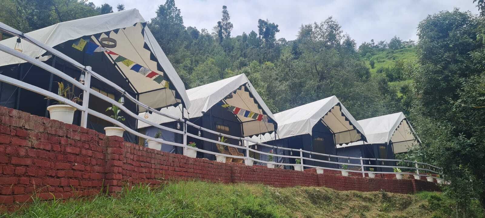 A row of traditional-style tents with white canopies, arranged in a linear fashion along a brick wall, set against a backdrop of lush greenery.