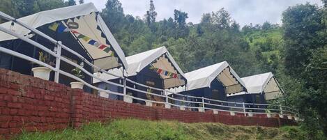 A row of traditional-style tents with white canopies, arranged in a linear fashion along a brick wall, set against a backdrop of lush greenery.
