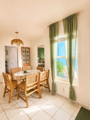 A dining area with a round table, wicker chairs, a wooden cabinet, and a window with green curtains.