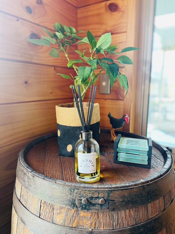 Un barril de madera con un difusor de botellas etiquetado "KINSHIP by ECOYA" y una pequeña planta en maceta con hojas verdes.