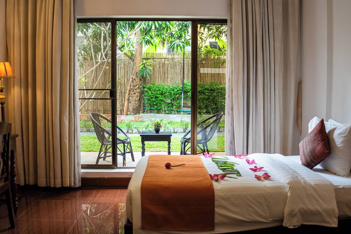 A hotel room with a bed, a view of a garden through a sliding door, and a small table with chairs outside.