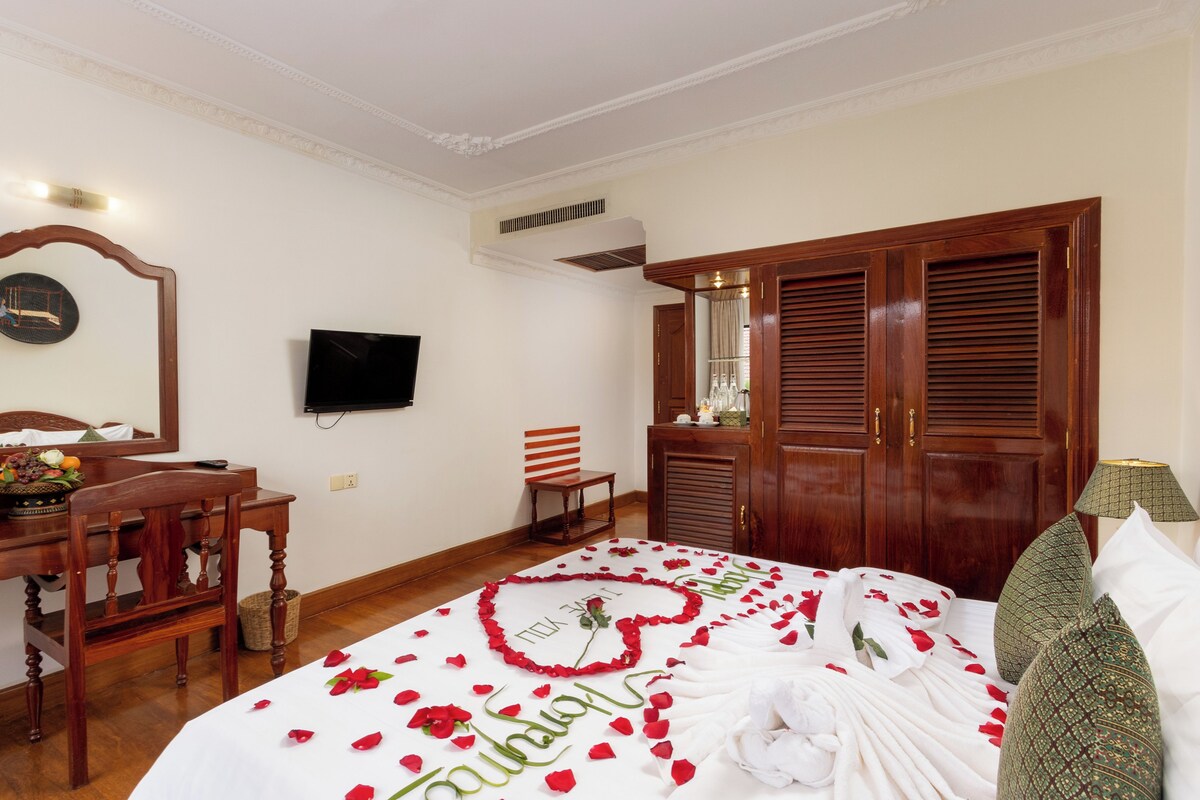 Habitación de hotel con una cama decorada con pétalos de rosa y un arreglo con forma de corazón, un escritorio de madera con una silla, un televisor montado en la pared y un amplio armario de madera.