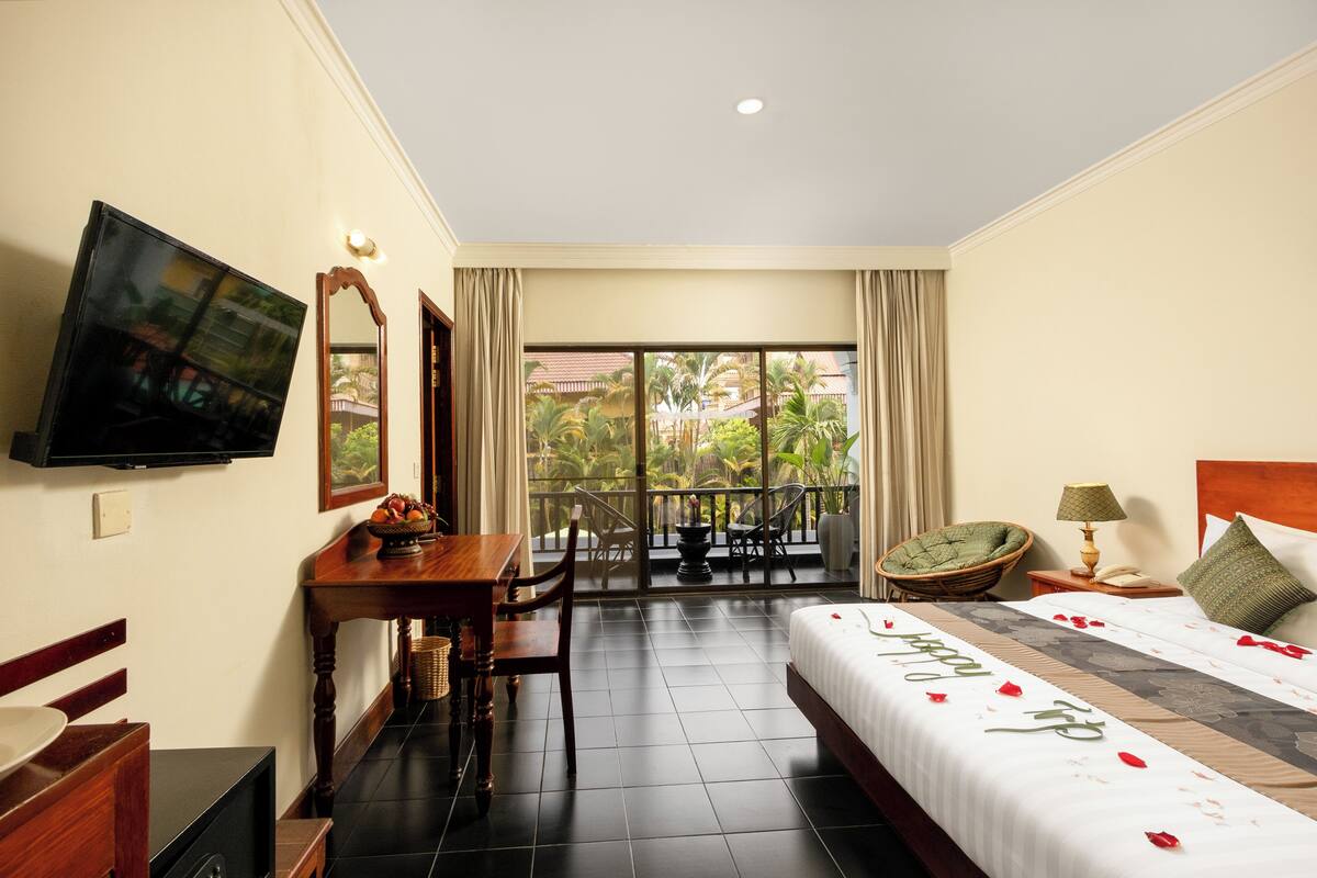 A hotel room with a bed, a desk with a chair, a television, a balcony with a view of palm trees, and a window with curtains.