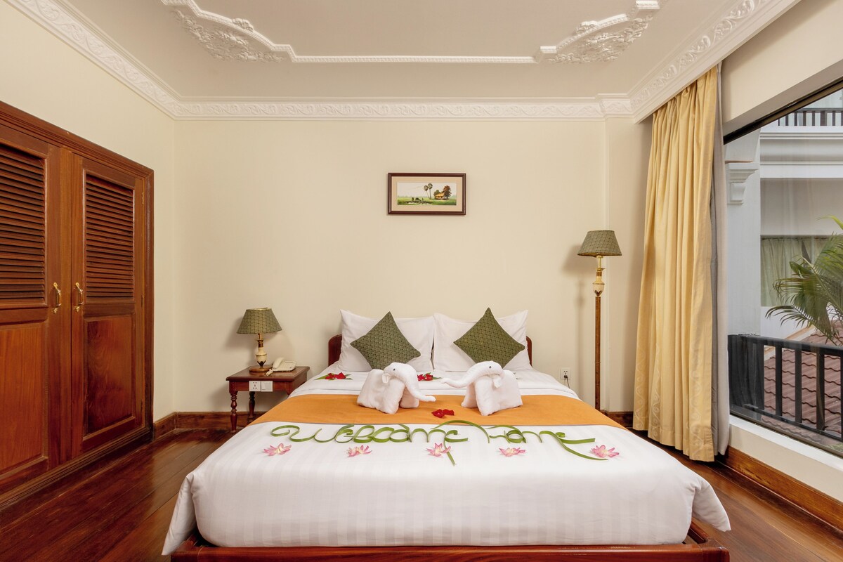 A hotel room with a large bed, wooden wardrobe, two bedside lamps, a framed picture on the wall, and a balcony view.